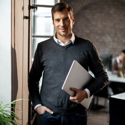 Portrait of smiling businessman holding laptop while standing in the office and looking at camera.